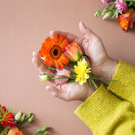 Hände halten einzelne Blüten ins Bild, unter anderem eine orangefarbene Gerbera