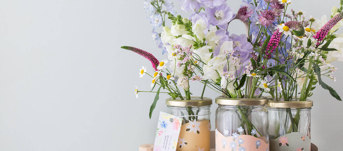 Mehrere mit Kreidefarbe angemalten Einmachgläser stehen auf einem Tablett und sind mit vielen schönen Blüten befüllt. Die Einmachgläser sind zudem dezent mit Blüten bemalt. An dem einen Einmachglas ist ein Zettel mit einer Grußnachricht befestigt.