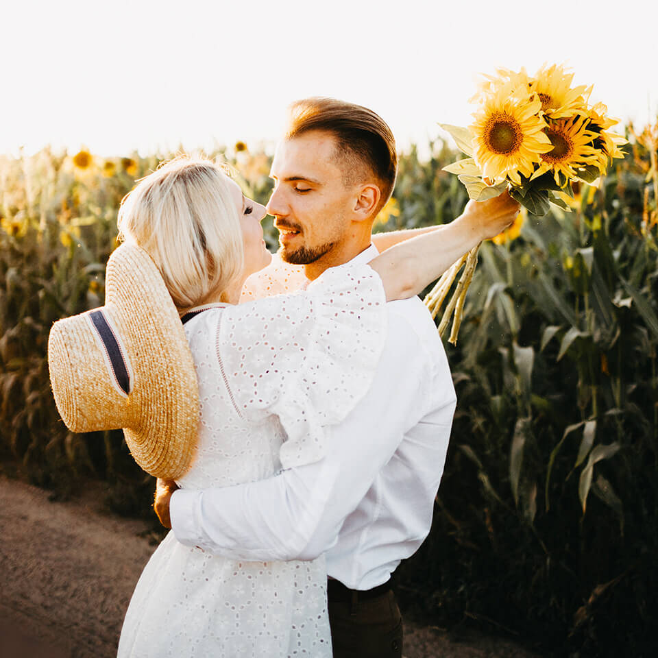 Ein Pärchen umarmt sich vor einem Sonnenblumenfeld. Die Frau hat ein paar Sonnenblumen in der Hand.