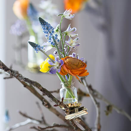 Zu sehen ist eine Mini-Vase, welche mit Frühblühern befüllt an einem kleinen Ast befestigt ist