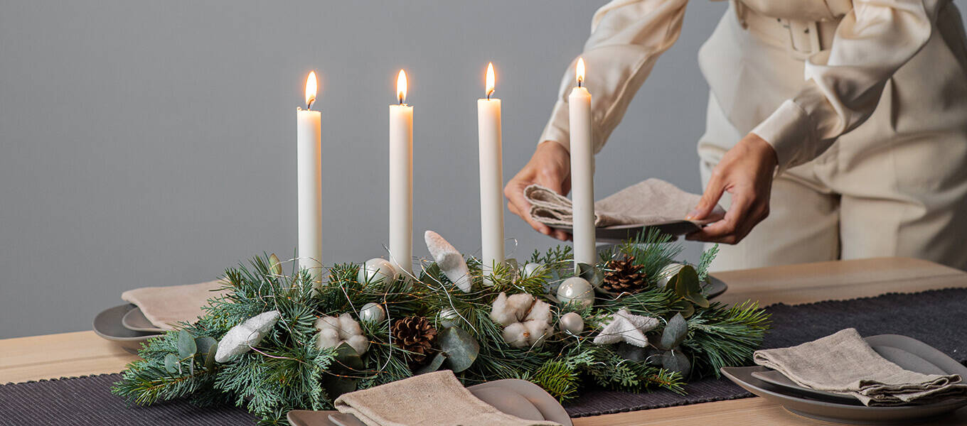 Modernes, längliches Adventsgesteck in grün weiß mit Stabkerzen. Eine Person dahinter dekoriert den Tisch weiter.